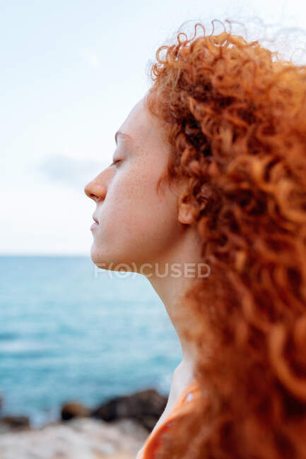 Спокойная женщина с вьющимися рыжими волосами наслаждается ветреной погодой на берегу бурлящего моря — стоковое фото