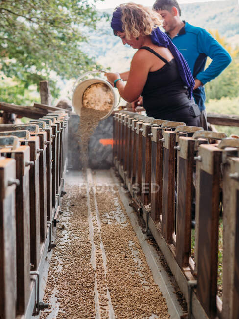 Vue latérale des agriculteurs mâles et femelles remplissant la nourriture du seau à la mangeoire debout près d'une clôture en bois dans la campagne montagneuse — Photo de stock