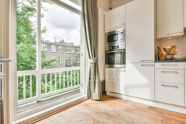 Innenraum der geräumigen Küche mit weißen Möbeln mit eingebauten Geräten in der Nähe von Balkonfenstern bei Tageslicht — Stockfoto
