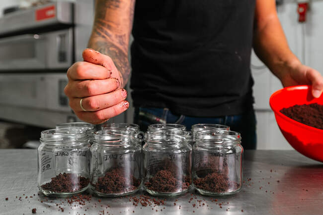 Crop male baker in casual outfit adding crumbs of chocolate cake into glass jars while preparing delicious layer dessert in kitchen — Stock Photo