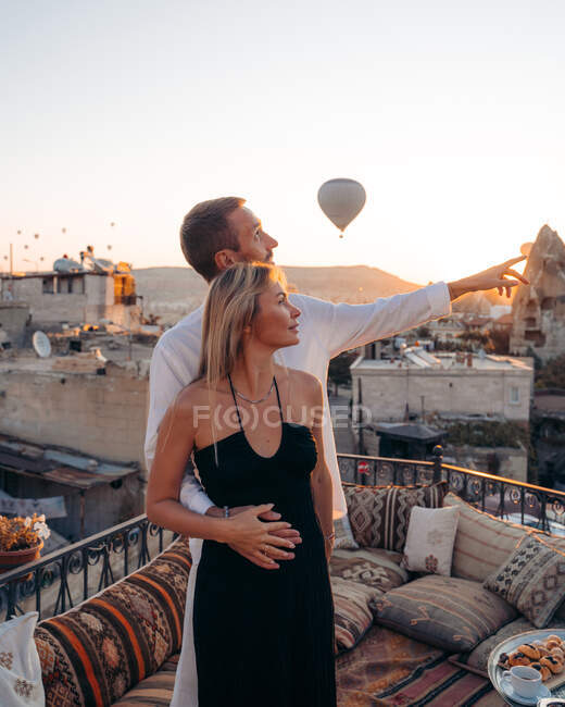 Hombre cariñoso abrazando a la mujer por detrás y apuntando hacia la terraza de la azotea con globos de aire caliente en el cielo de la noche en Capadocia Turquía - foto de stock