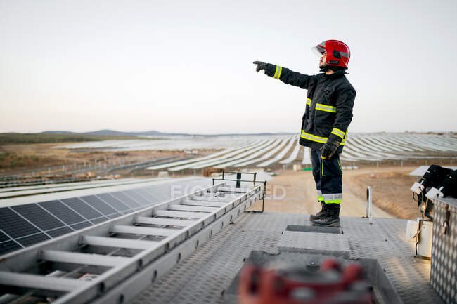 Full body side view of male firefighter wearing uniform and gloves standing on metal roof of fire engine near ladder with hand pointing forward at solar panels in rural farm — Stock Photo
