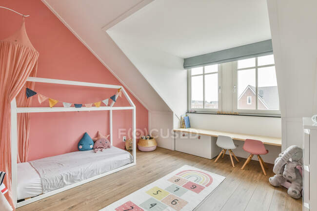 Comodo letto a baldacchino con giocattoli situato vicino alla finestra e scrivania in soffitta moderna vivaio con pareti bianche e rosa — Foto stock