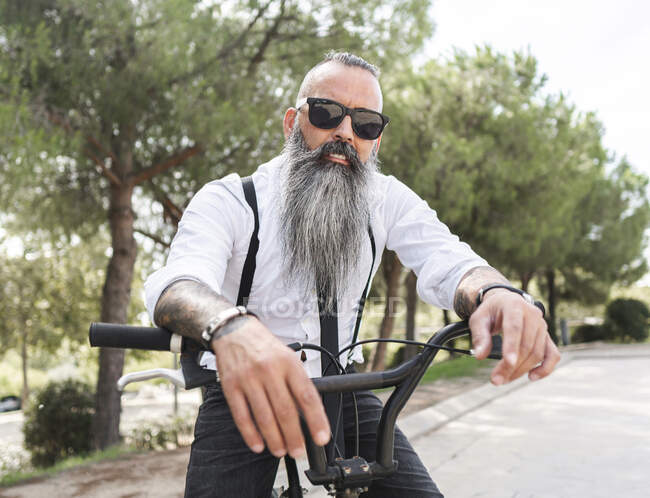 Confiado hipster masculino con tatuajes en camisa blanca y gafas de sol sentado en bicicleta en el parque con árboles verdes en la ciudad - foto de stock