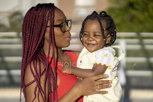 Cheerful African American mother with red braids standing with positive little daughter on hands on street in sunlight — Stock Photo