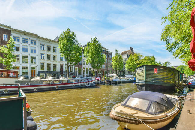 Sporco fiume increspato con barca a motore ormeggiato e navi contro facciata edificio multipiano sotto cielo nuvoloso nel porto di Amsterdam Paesi Bassi — Foto stock