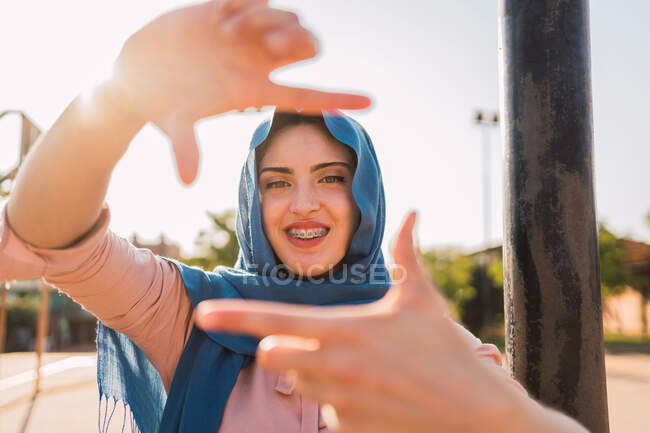 Mujer árabe feliz en hijab mostrando el signo de encuadre y mirando a la cámara mientras está de pie en el día soleado en la calle - foto de stock