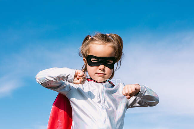 D'en bas petite fille en costume de super-héros levant les poings tendus pour montrer la puissance tout en se tenant contre le ciel bleu clair — Photo de stock