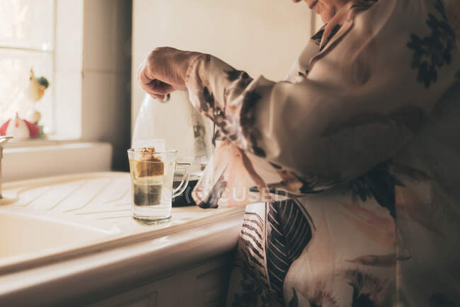 Cropped unrecognizable adult female wearing silk blouse and trousers brewing tea bag in glass mug kitchen — Stock Photo