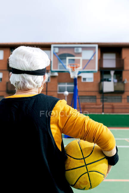 Анонимный спортсмен в спортивной одежде, стоящий на площадке с желтым мячом и баскетбольным кольцом во время игры на улице — стоковое фото