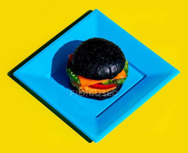 Dall'alto hamburger fresco con panino nero e verdure servite su piatto quadrato blu su sfondo giallo — Foto stock