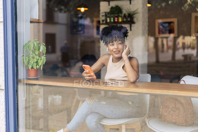 Through window of young ethnic female with dark Afro hair in trendy outfit and true wireless earphones happily while having video conversation on smartphone in modern cafe — Stock Photo