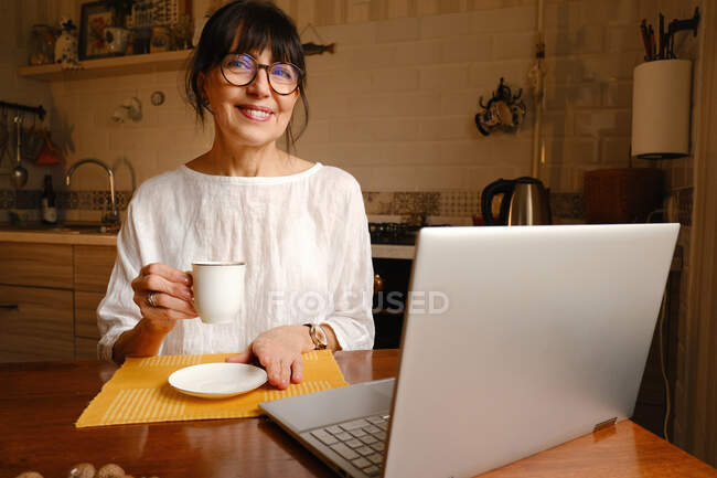 Mujer positiva en anteojos sentada en la mesa con taza de bebida caliente y portátil en la cocina por la mañana y mirando a la cámara - foto de stock