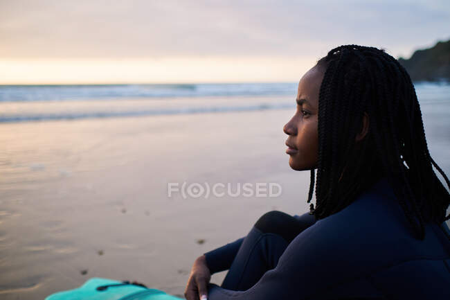 Vista lateral de la mujer afroamericana pensativa con pelo rizado corto en traje de neopreno sentado en la playa de arena cerca del océano y admirando la puesta de sol - foto de stock