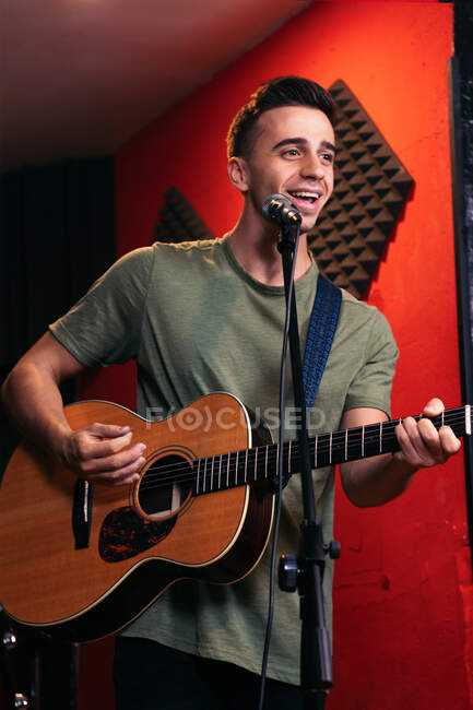 Positive young male guitarist playing acoustic guitar and singing in microphone in light club — Stock Photo