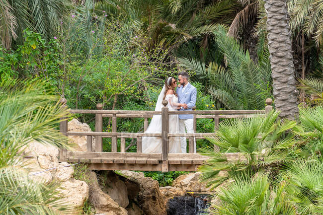 Newlywed couple in wedding outfits standing on wooden footbridge with railing and holding hands while hugging and looking at each other above waterfall with rocks near green trees in park — Stock Photo