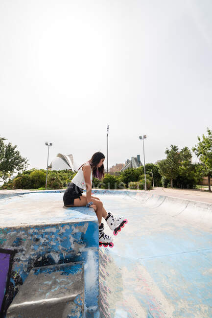 Vista laterale del corpo pieno giovane donna che indossa top bianco e pantaloncini jeans neri in pale a rulli bianchi seduti sulla rampa di strada in skate park — Foto stock