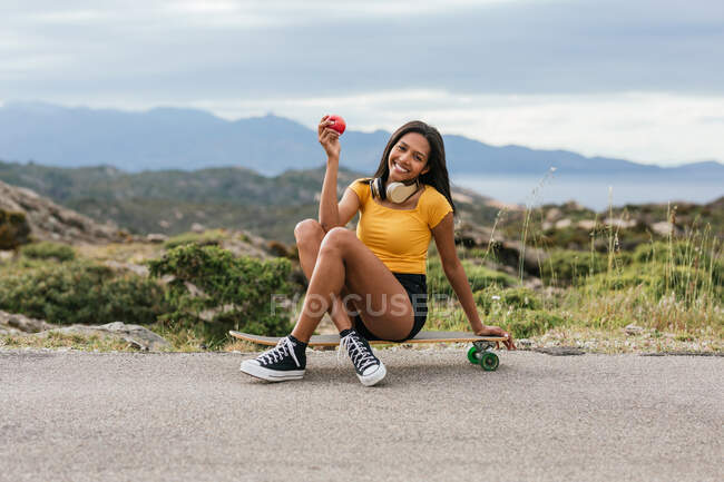 Corpo pieno di femmina etnica positiva con cuffie wireless sul collo e mela in mano mentre seduto sul longboard su strada asfaltata — Foto stock