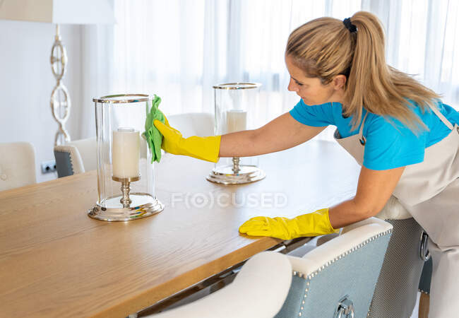 Vista lateral de la joven limpiadora profesional concentrada en uniforme y guantes limpiando el polvo del elegante candelabro colocado en el comedor en la espaciosa casa - foto de stock