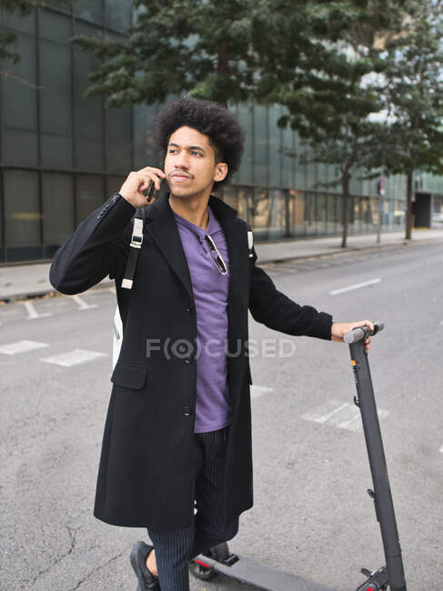 Full body of confident young African American male millennial with dark curly hair in stylish outfit talking on smartphone while standing on city street with electric scooter — Stock Photo
