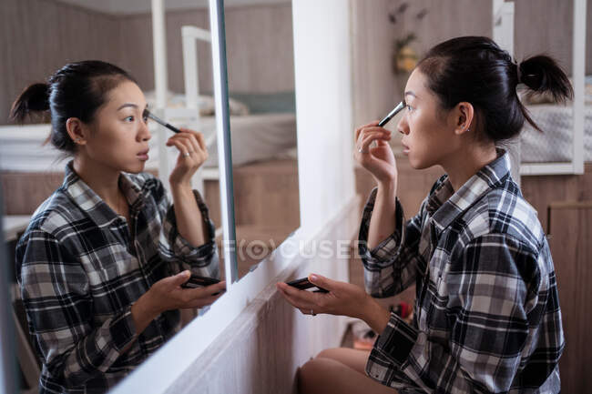 Focalizzato affascinante femmina etnica applicando ombretto con pennello durante il trucco e guardando nello specchio — Foto stock