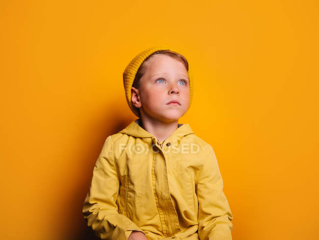 Niño poco emocional en impermeable de moda y gorro sombrero de pie mirando hacia otro lado contra el fondo amarillo en el estudio - foto de stock