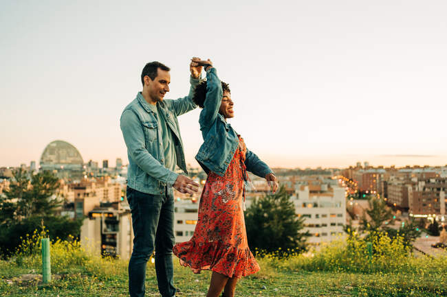 Feliz joven pareja diversa sosteniendo la mano mientras bailan juntos en el prado floreciente en el día soleado - foto de stock
