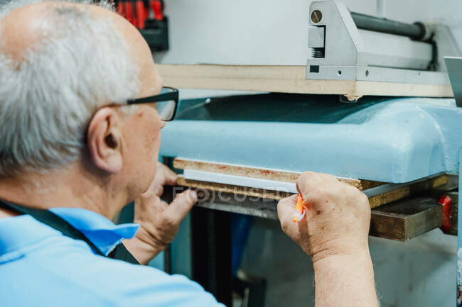 Crop elderly male worker in casual clothes and eyeglasses making marks on pile of paper sheets while operating printing press machine — Stock Photo