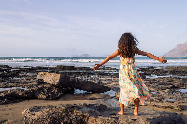 Back view of anonymous little girl tourist in sundress with outstretched arms contemplating sea from Famara Beach in Canary Islands — Stock Photo