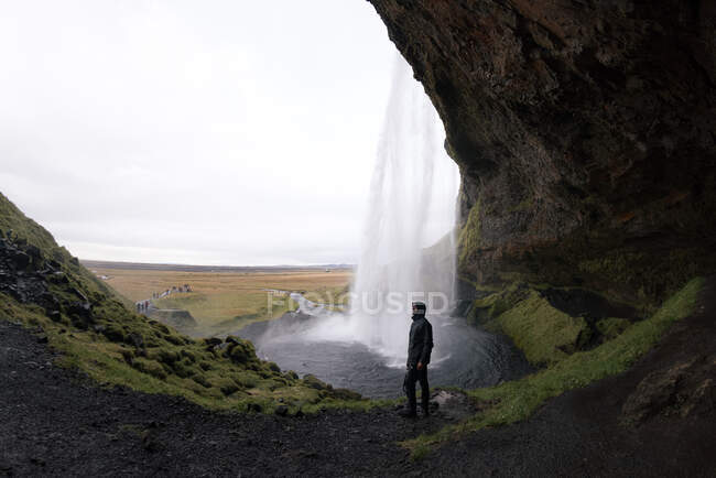 Вид сбоку на анонимного путешественника в теплой одежде, стоящего в скалистой пещере и любующегося живописным быстрым водопадом Сельджаландсфосс под облачным небом в Исландии — стоковое фото