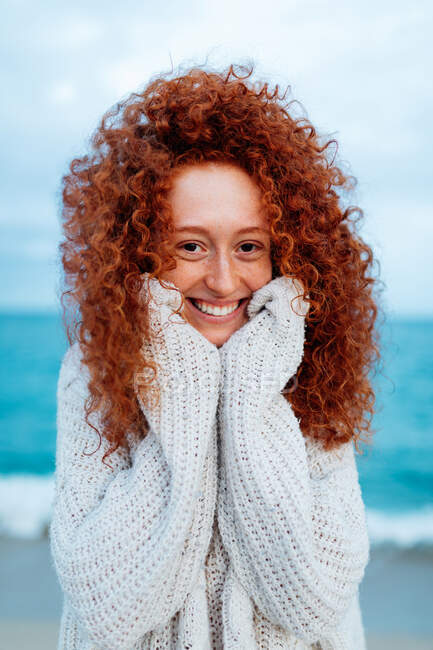 Восхитительная женщина с кудрявыми длинными рыжими волосами в трикотажном свитере, стоящая перед камерой на фоне синего моря — стоковое фото