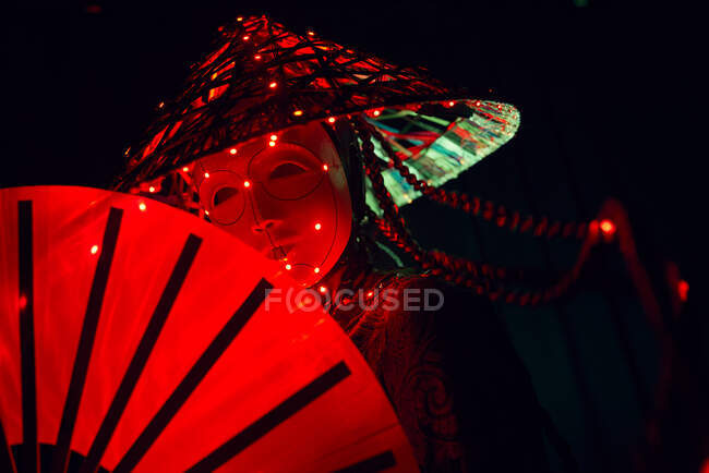 Unrecognizable female with a mask in creative traditional outfit and Vietnamese headwear with red illumination standing in dark studio on black background during performance — Stock Photo