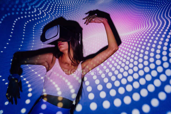 Femme à la mode dans le crop top expérience de la réalité virtuelle dans le casque tout en dansant dans les projecteurs lumières — Photo de stock
