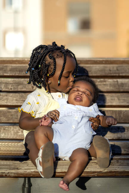 Спокійна афроамериканська дівчинка з плечима в повсякденному одязі сидячи з дитиною на дерев'яній лавці на вулиці в сонячний день. — стокове фото