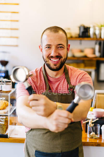 Positivo barista maschio in grembiule guardando la fotocamera e mostrando portafiltri speciali mentre in piedi vicino al bancone nella moderna caffetteria — Foto stock
