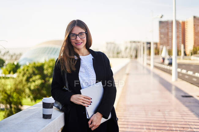 Позитивная молодая предпринимательница в черном пальто, стоящая с нетбуком и чашкой горячего напитка на вынос и широко улыбающаяся — стоковое фото