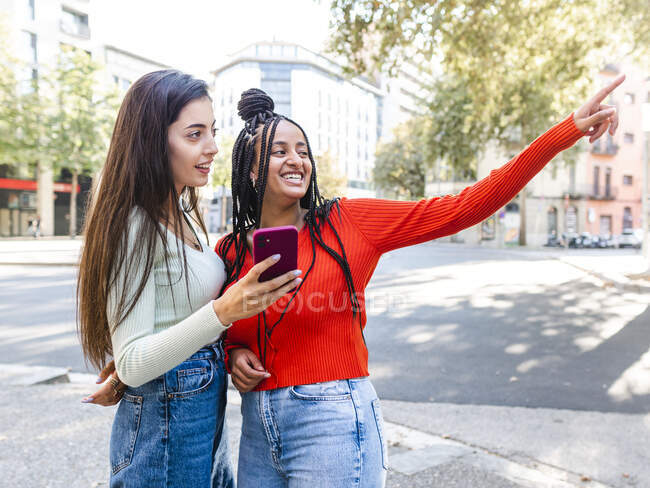 Seitenansicht fröhlicher Frauen in stylischer Kleidung, die auf der Straße stehen und lachen, während sie tagsüber auf dem Smartphone surfen — Stockfoto