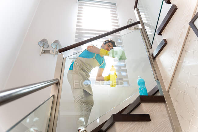 Снизу положительный работник мужского пола в фартуке и резиновых перчатках улыбается, очищая стекло лестницы тканью — стоковое фото