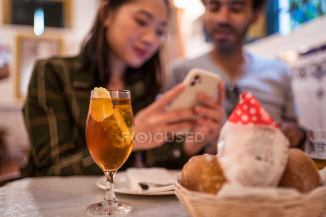 Cosecha joven mujer asiática mostrando fotos en el teléfono inteligente a novio étnico sentado en la mesa con vaso de limonada y cesta de pan en el restaurante - foto de stock