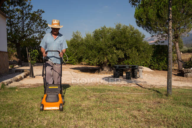 Pieno corpo di giardiniere maschio irriconoscibile in cappello falciare prato erboso vicino a cespugli e alberi in estate — Foto stock