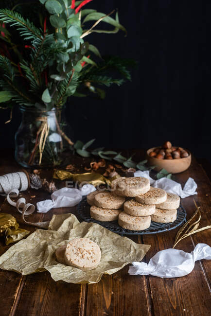 Куча аппетитных сладких песочных печенек с фундуком на тарелке на деревянном столе с праздничной оберточной бумагой и лентами для празднования Рождества — стоковое фото