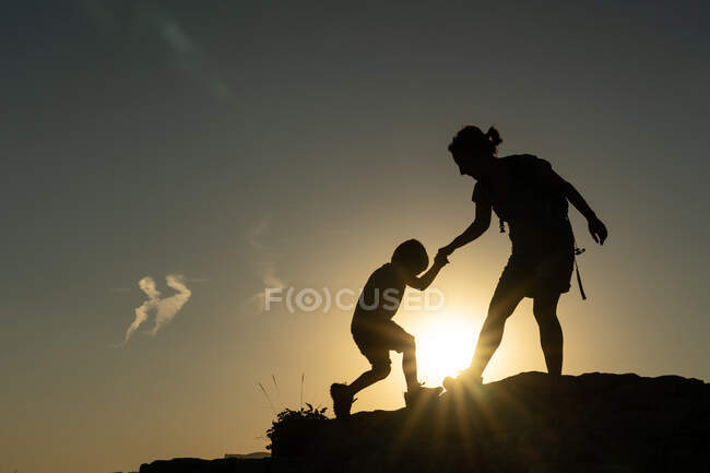 Mãe ajudando seu filho a subir uma montanha contra a luz silhueta pelo sol em uma história motivacional — Fotografia de Stock