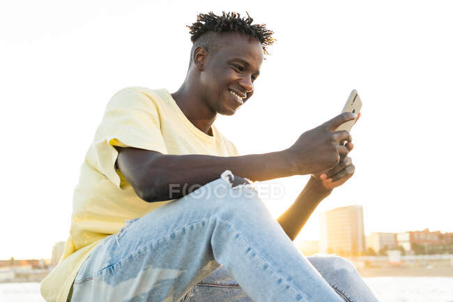 Улыбающийся афроамериканец в повседневной одежде сидит на скалистом побережье, используя смартфон в летний вечер — стоковое фото