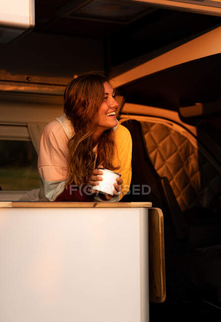 Entzückte Forscherin mit einem Becher heißen Tees steht im Reisemobil im Sonnenuntergang und genießt den Abend in Spanien, während sie wegschaut — Stockfoto