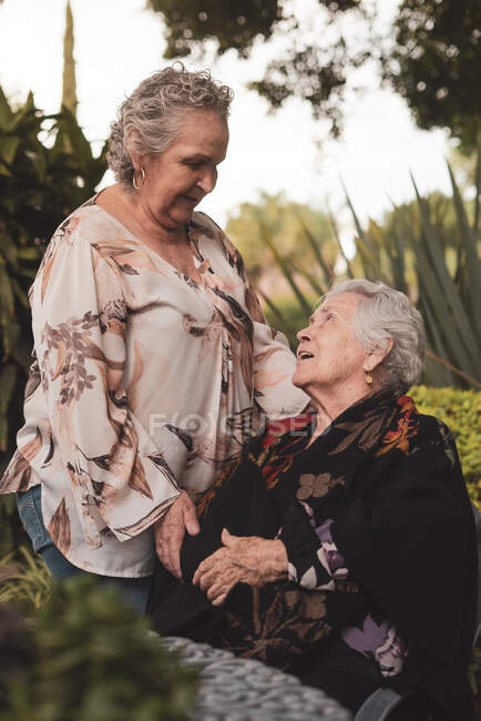 Feliz anciana abrazando a la hermana en la cabeza y luego sonriendo mirándose mientras pasan tiempo juntos en el jardín - foto de stock