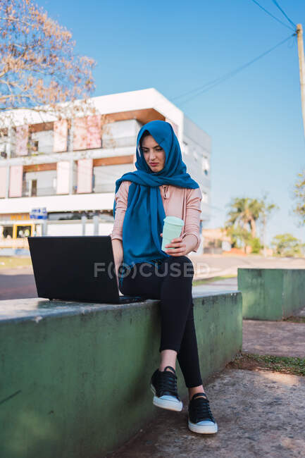 Улыбающаяся арабская фрилансер в хиджабе сидит на скамейке с кофе на вынос и просматривает нетбук во время работы над проектом удаленно — стоковое фото