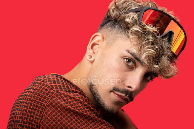 Side view of cool male model with curly hair and modern sunglasses looking at camera against red background — Stock Photo