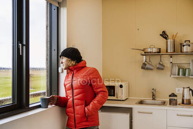 Calmo jovem viajante masculino em casaco vermelho quente e chapéu bebendo xícara de café quente e olhando para fora janela de pé na cozinha em casa no dia chuvoso no campo — Fotografia de Stock