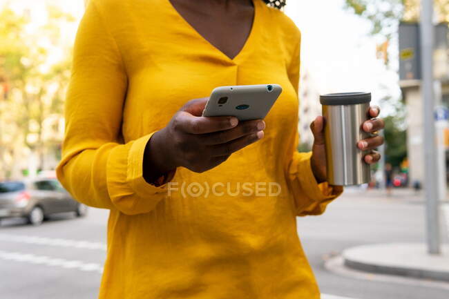 Mulher afro-americana anônima com caneca térmica enquanto mensagens de texto no celular na rua na cidade — Fotografia de Stock