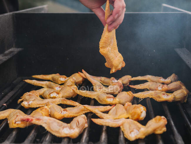 Crop chef anónimo poniendo ala de pollo crudo en la parrilla de metal caliente con humo mientras se cocina en el campo durante el picnic - foto de stock